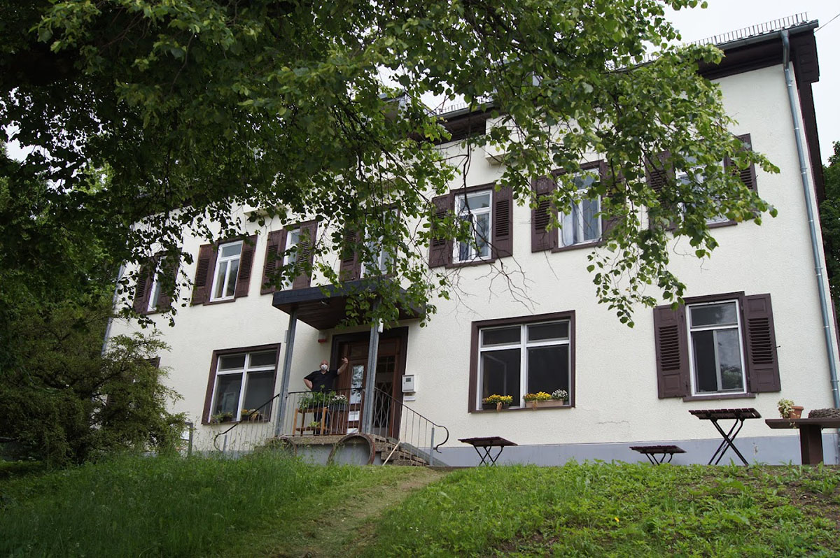 Zentrales Gebäude des Waldeckhofs: ein zweigeschossiges, hell verputztes Haus mit vielen rechteckigen Fenstern und dunkelbraunen Fensterläden. Vor dem Eingang befindet sich eine kleine überdachte Veranda mit Treppe, Geländer und einigen Pflanzen. Auf der Veranda steht eine Person. Das Gebäude ist von grünen Bäumen umgeben, deren Äste teilweise die Ansicht verdecken. Im Vordergrund liegt ein leicht ansteigender Grasbereich mit einem schmalen Trampelpfad sowie einigen einfachen Holzbänken und kleinen Tischen.